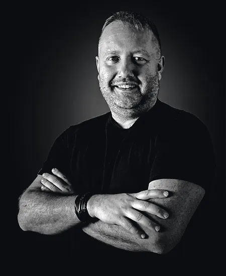 Dimitri photographed in black and white against a dark background, arms folded, close-cropped hair and a short beard, in a plain dark t-shirt with a braided band on one wrist, smiling at the camera